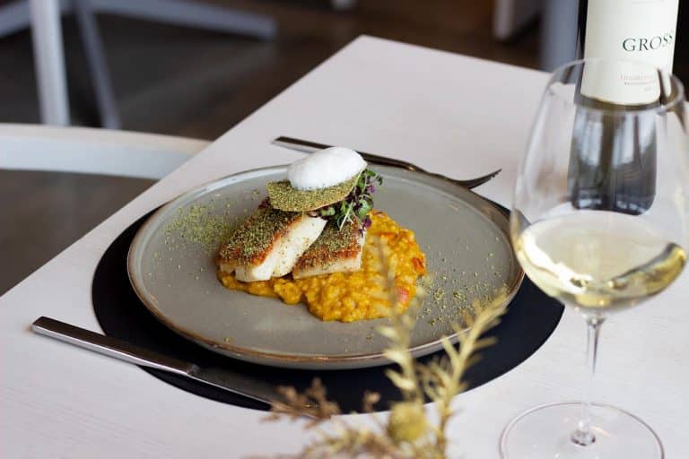 Ein kunstvoll angerichtetes Fischgericht auf einem grauen Teller. Der Fisch ist knusprig gebraten und wird auf einem Bett aus cremigem Kürbisrisotto serviert. Im Hintergrund steht ein Glas Weißwein sowie eine Flasche Wein, die teilweise sichtbar ist.